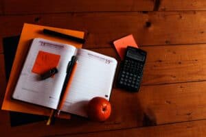 office supplies on a dark wood desk including calendar, sticky notes, pen, orange folder, and a mandarin for a snack