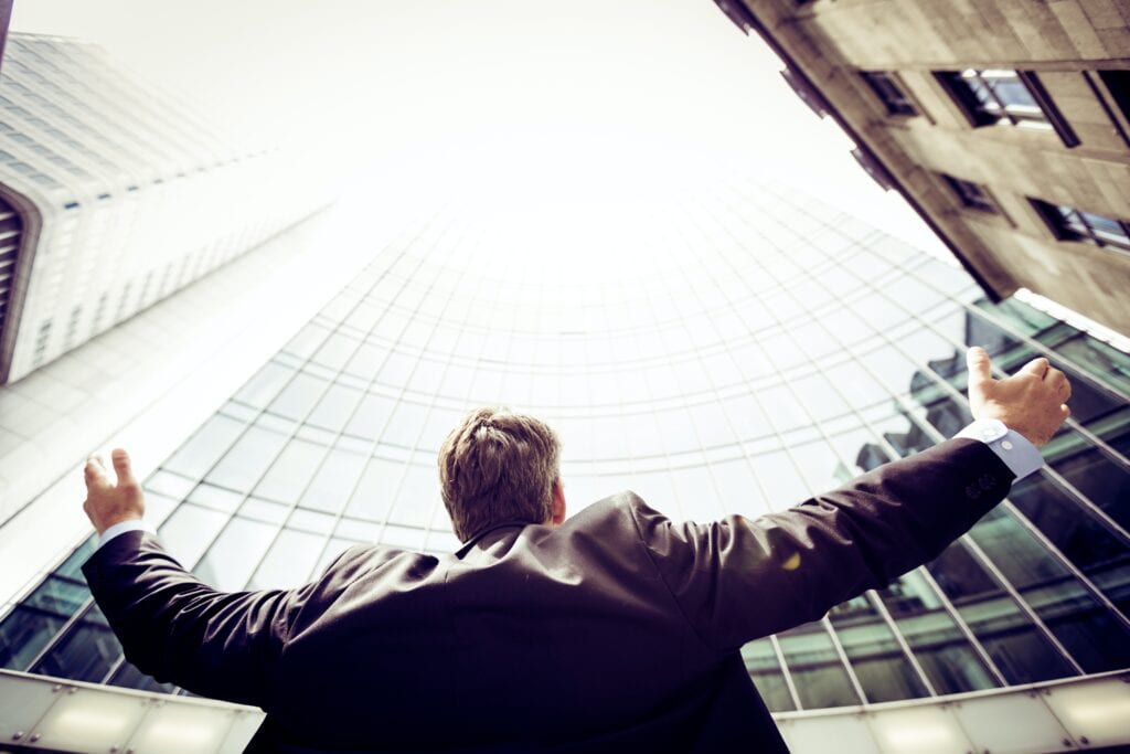 man in city downtown looking up at a building of windows arms outstretched celebrating sales hiring roi