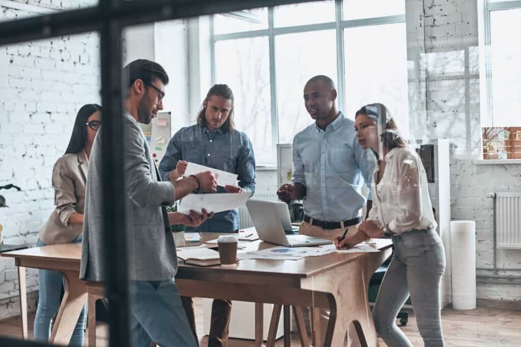 Analyzing sales. Group of young modern people communicating together while working behind the glass wall in the board room empowered by effective coaching leadership.