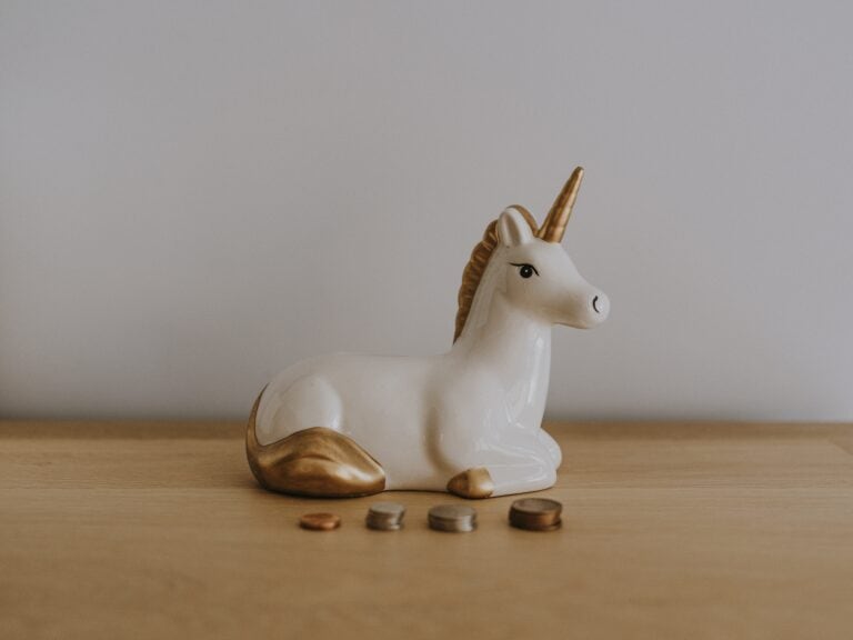 White ceramic unicorn with gold horn lying down in front of small stacks of coins