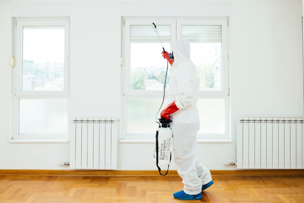 pest control specialist in all white room, wearing white coveralls with red gloves spraying pest spray also does sales prospecting on site