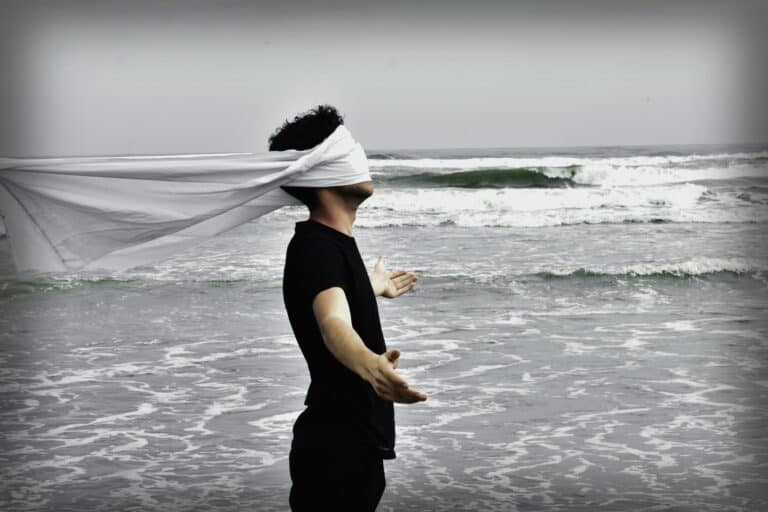 man in black standing at the ocean with arms widespread and blindfolded
