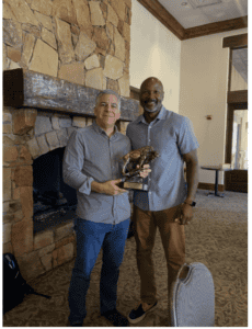 2 men standing by fireplace. one many with buffalo statue award trophy