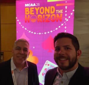 Selfie image of two men in front pink and orange signage at the annual MCAA conference
