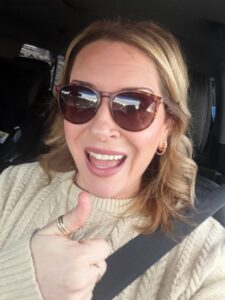 blonde female in car with seatbelt on. cream sweater, brown sunglasses, giving a thumbs up.