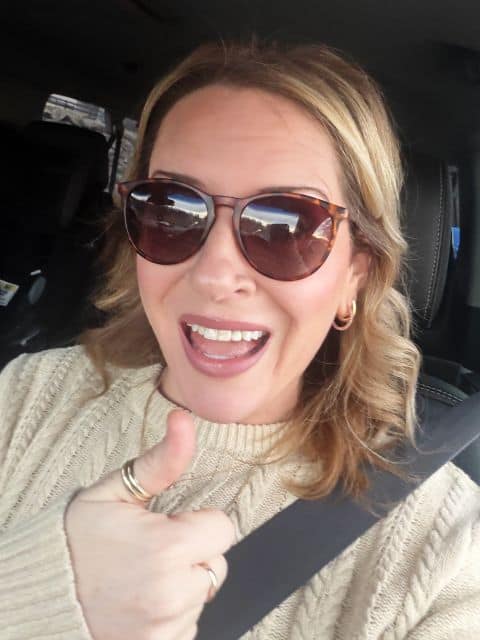 blonde female in car with seatbelt on. cream sweater, brown sunglasses, giving a thumbs up.