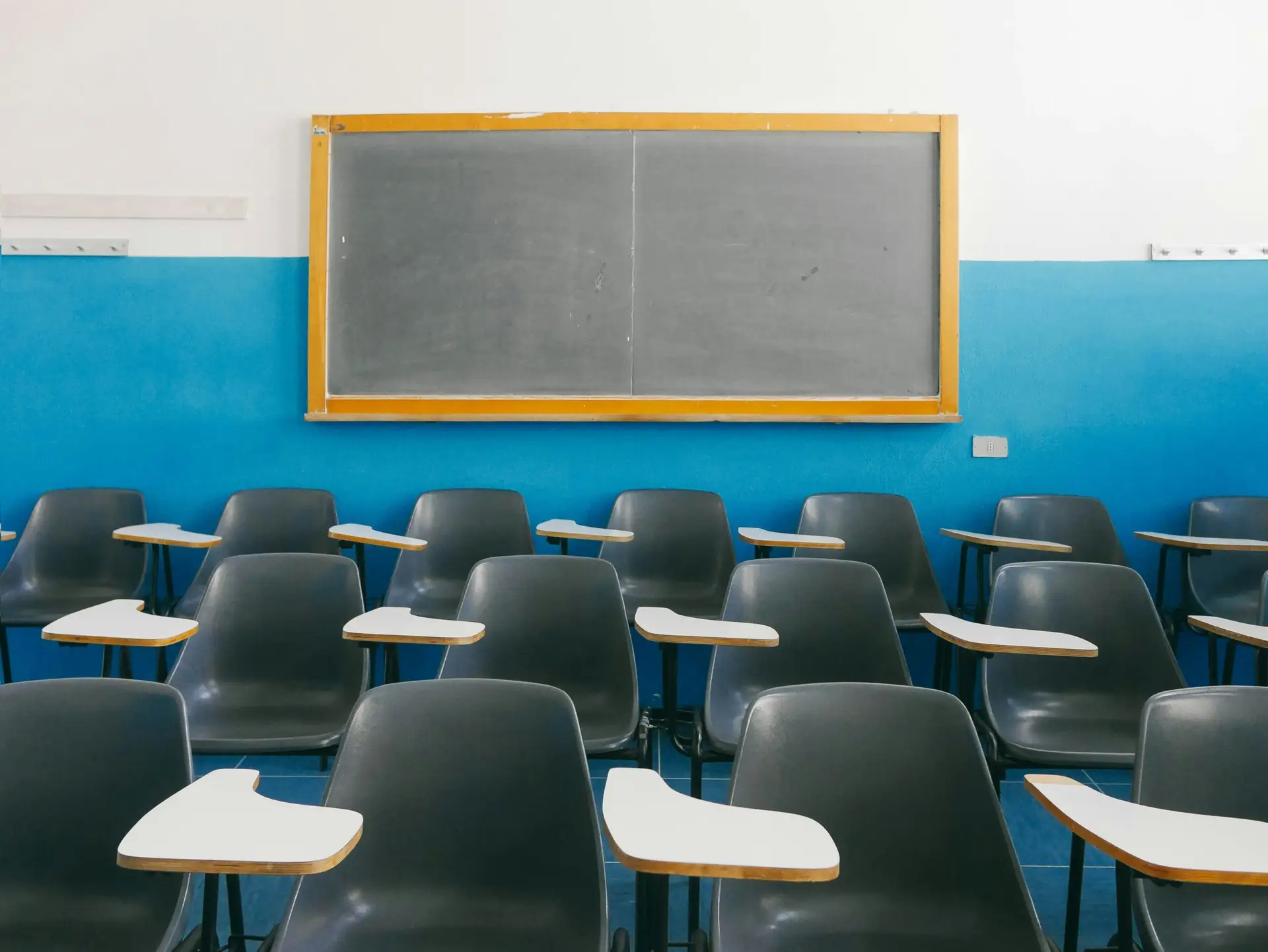 Empty classroom full of black chairs with side desks with a clean chalkboard against a blue and white wall in the background